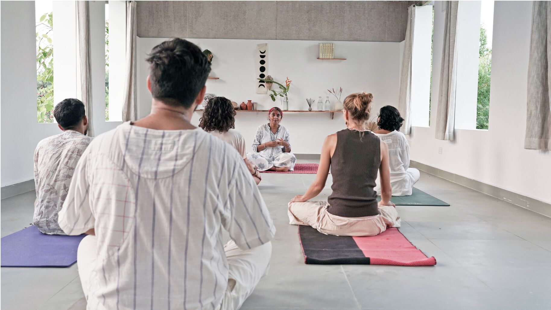A group of people sitting on yoga mats in a room with large windows and a neutral color scheme.