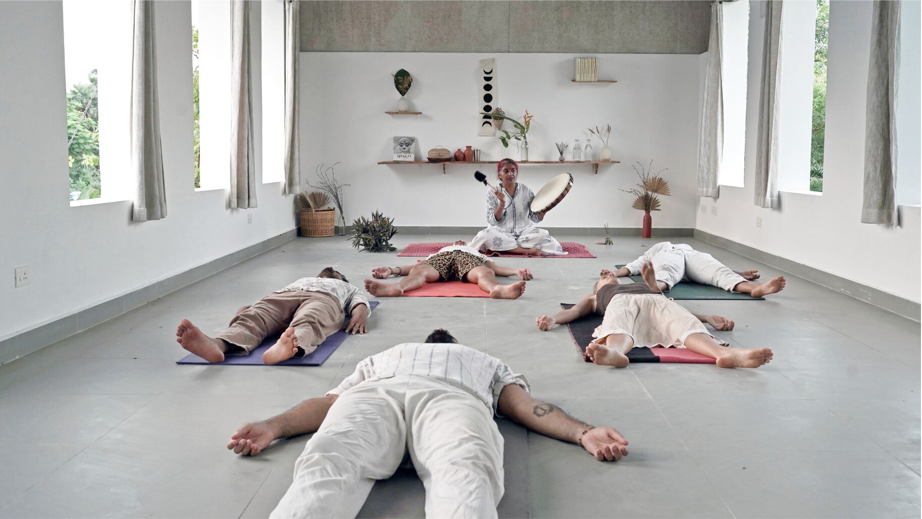 A spacious room with large windows, a yoga mat area, and a wall decorated with plants and shelves.