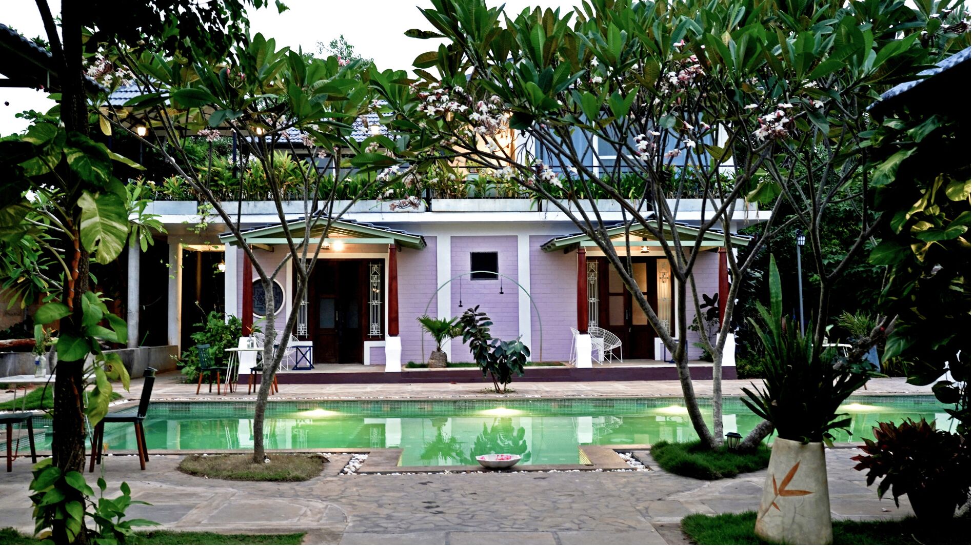 A two-story building with a pool in the foreground, surrounded by lush greenery.