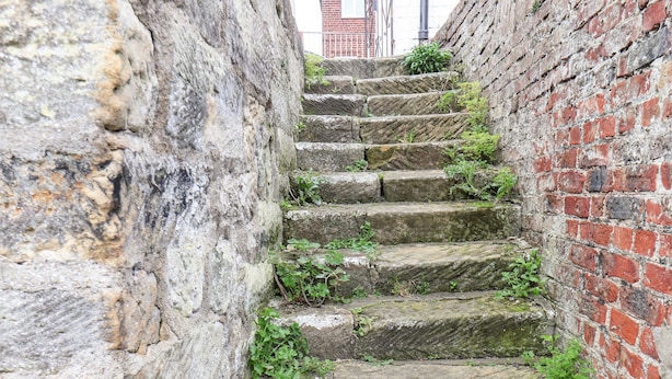 Una estrecha escalera de piedra con escalones que ascienden, flanqueada por un muro de piedra a un lado y un muro de ladrillo al otro.