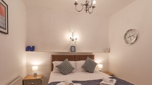 Un dormitorio ordenado con una cama grande, dos mesitas de noche, un revistero, un reloj de pared y una araña.