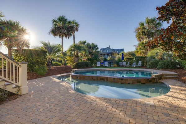 A swimming pool with a brick patio, surrounded by palm trees and a house in the background.