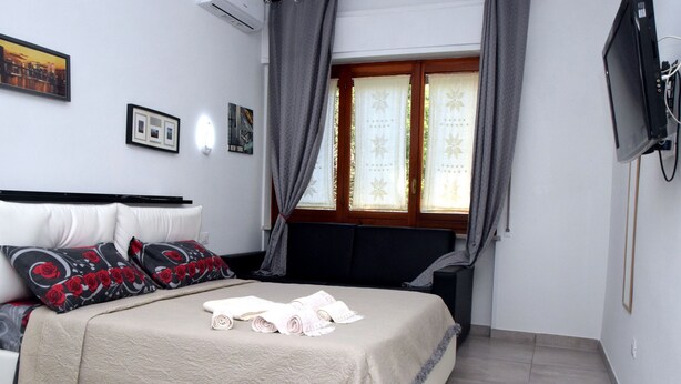 A neatly made bed with a beige cover, a black headboard, and red and black patterned pillows. There are two framed pictures on the wall and a television mounted on the wall. - B&B Royal Reggia di Caserta (Casagiove)