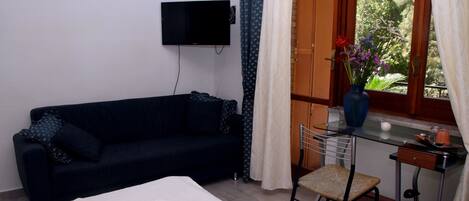 Una habitación con cama, un sofá azul oscuro, un escritorio pequeño con una silla, un televisor montado en la pared y una ventana con cortinas.