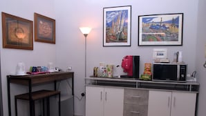 A kitchenette with a microwave, coffee maker, and various food items on the counter. There is a small table with a chair and a lamp. Two framed pictures are hung on the wall above the counter. - B&B Royal Reggia di Caserta (Casagiove)