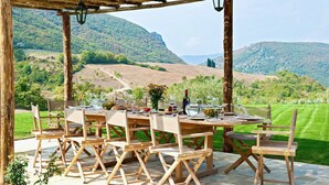 Un área de comedor al aire libre con una pérgola de madera, mesas dispuestas para una comida y una vista panorámica de colinas ondulantes y viñedos.