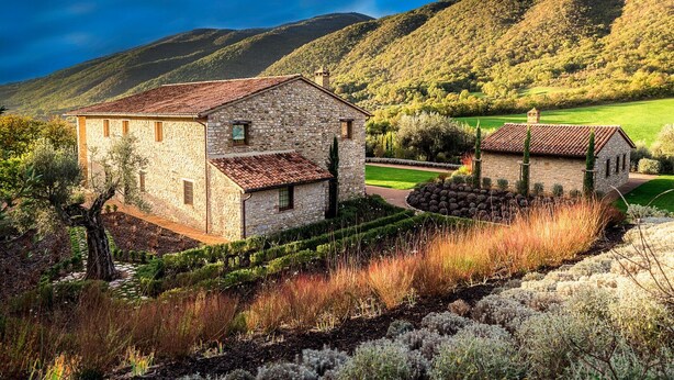 Una casa de piedra con tejado de baldosas, rodeada de un jardín cuidado y con vistas a colinas ondulantes al fondo.