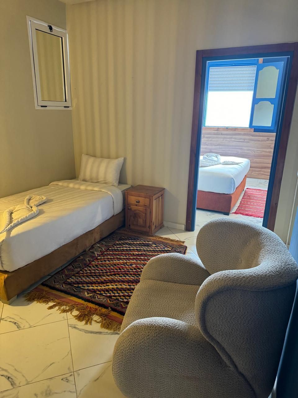 Une chambre avec un lit, une table de chevet en bois, une chaise, un tapis à motifs et une vue sur une autre pièce par une porte ouverte.