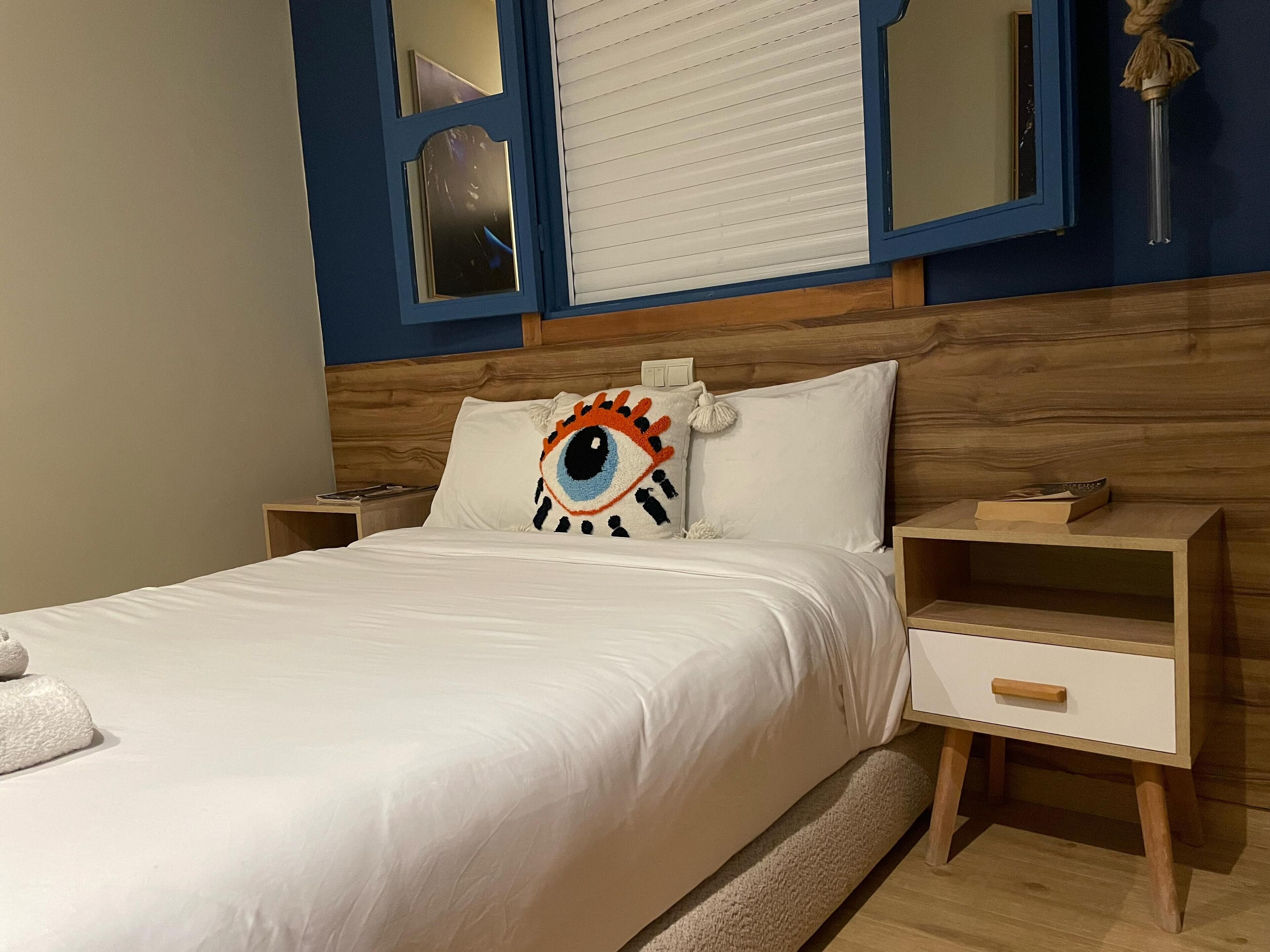 A neatly made bed with a white comforter and pillows, a wooden headboard, a bedside table with a book, and a nightstand with a lamp.