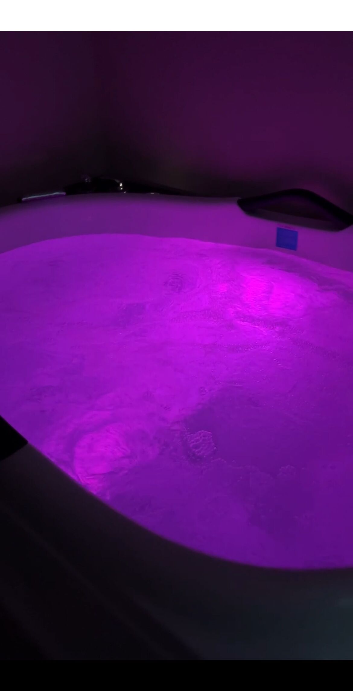 Ein Raum mit einem violett beleuchteten, kreisförmigen Bereich, möglicherweise ein Pool oder eine Whirlpool, und einem blauen Gegenstand an der Wand.
