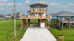 A two-story wooden structure with a balcony, a garage, and a gravel driveway. - 7 Day Weekend: 4  BR, 3  BA House in Crystal Beach, Sleeps 17 (Bolivar Peninsula)
