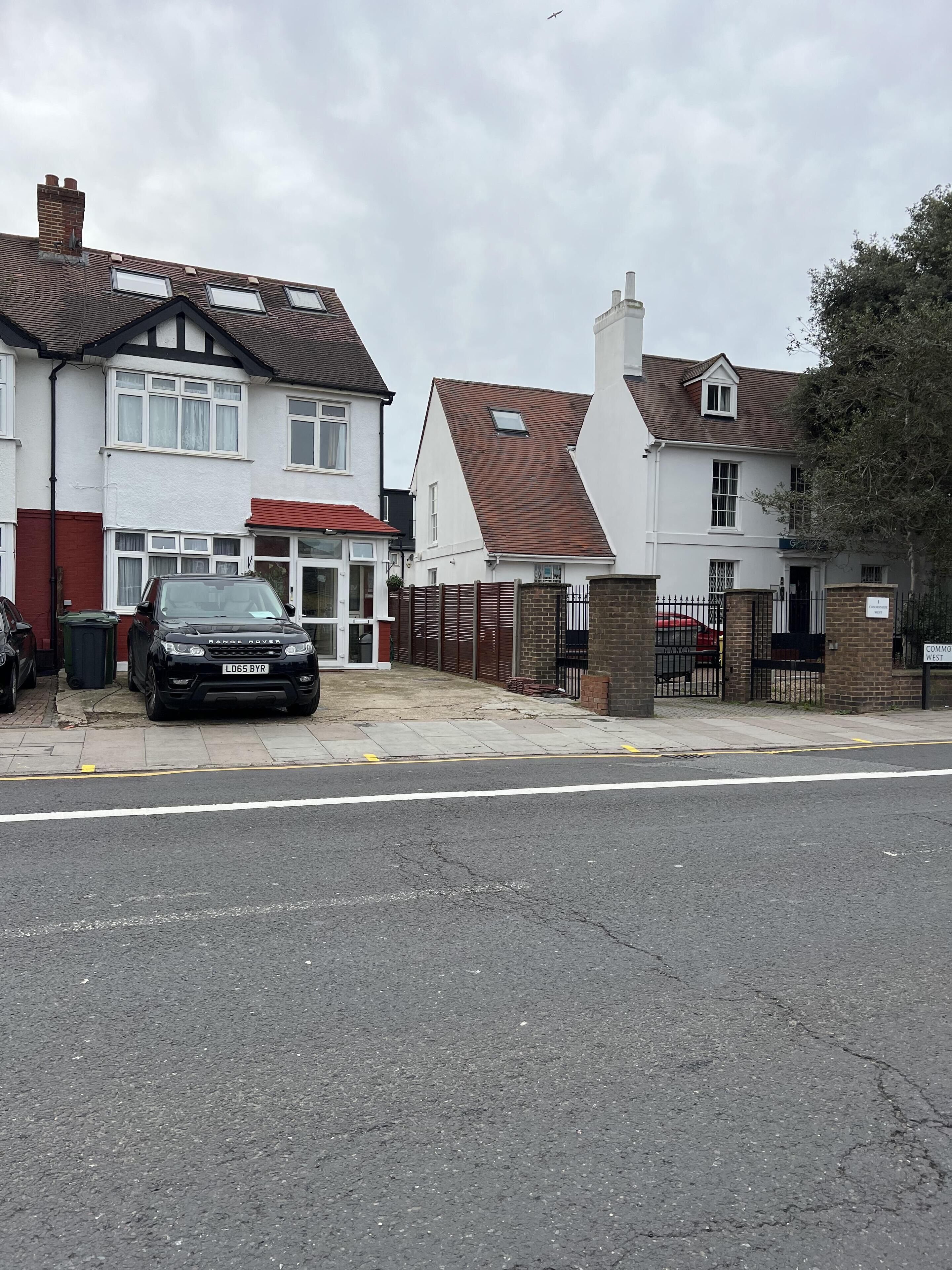 A residential street with houses, a black car parked on the side, and a gated entrance.