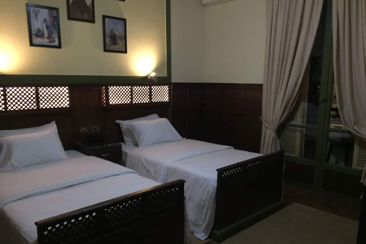 Quarto com duas camas de solteiro, uma mesa de cabeceira, luminária na parede e janela com cortinas.