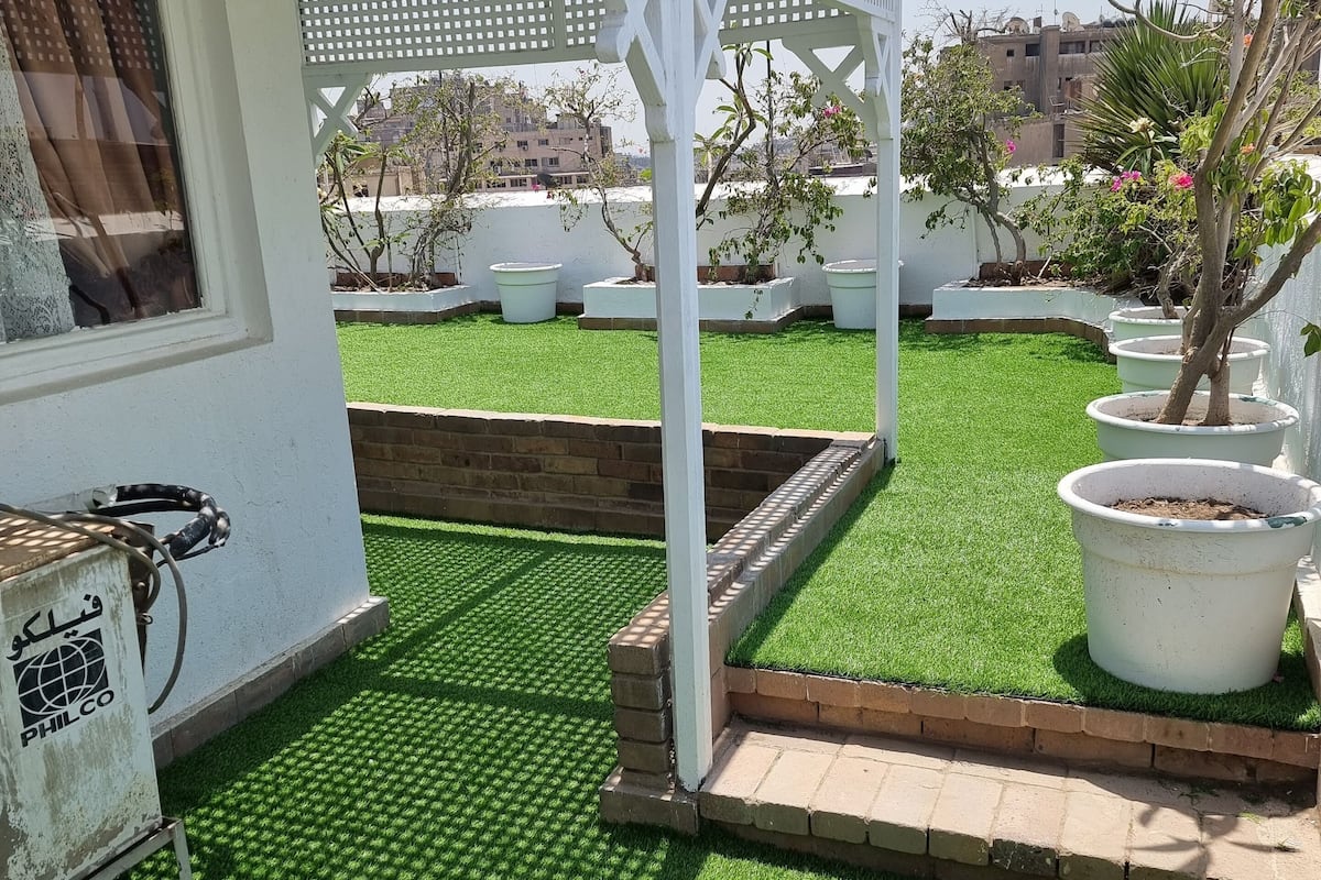 Um edifício branco com um alpendre coberto, grama artificial verde e plantas em vasos.
