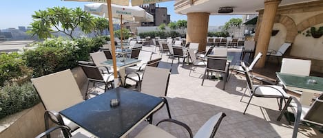 An outdoor café with tables and chairs, large umbrellas, and a view of the cityscape.