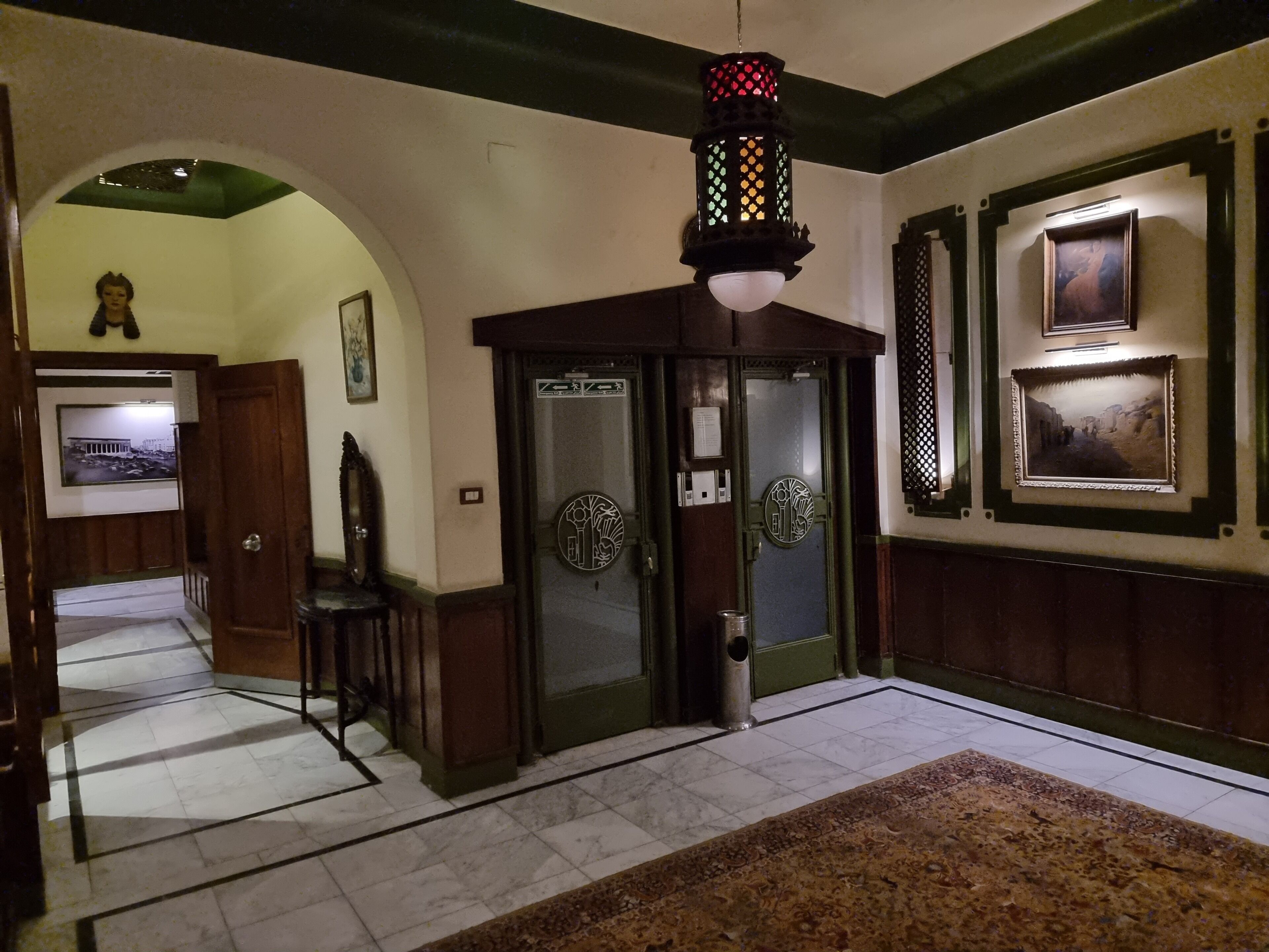 A lobby with a tiled floor, a patterned rug, a wooden bench, a door with a green frame, and a hanging lantern.