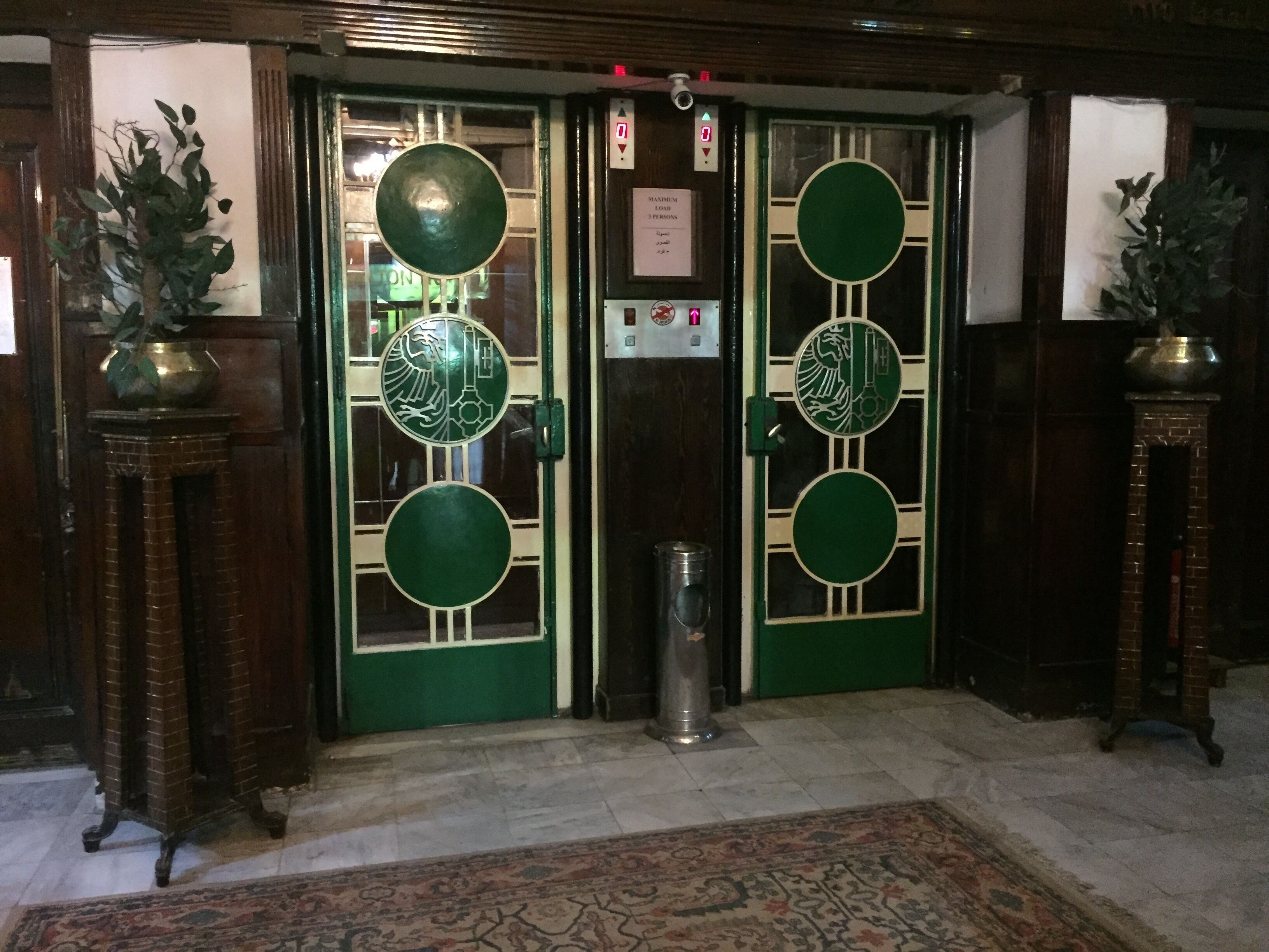 A set of double doors with intricate designs, a brass door knocker, and a small trash bin in front.