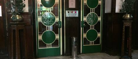 A set of double doors with intricate designs, a brass door knocker, and a small trash bin in front.