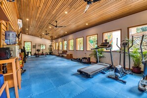 A gym with exercise equipment, a blue rubber floor, wooden ceiling, and large windows.