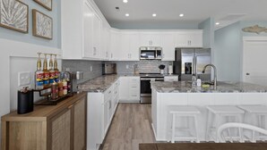 Une cuisine moderne avec des meubles blancs, un plan de travail en granit, des appareils électroménagers en acier inoxydable et un coin bar avec des bouteilles d’alcool.