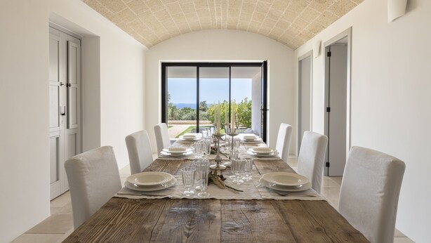 A dining area with a long wooden table set for six, white chairs, and a view of the sea through large windows. - Finamore - Panoramic villa with sea view, pool, forest, 4-5 bedrooms, Monopoli (Cozzana)