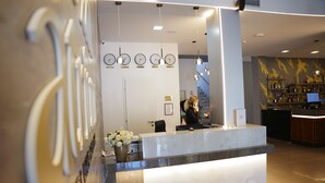 Zona de recepción de un hotel con un mostrador de mármol, un escritorio de recepción, un reloj y una escalera al fondo.