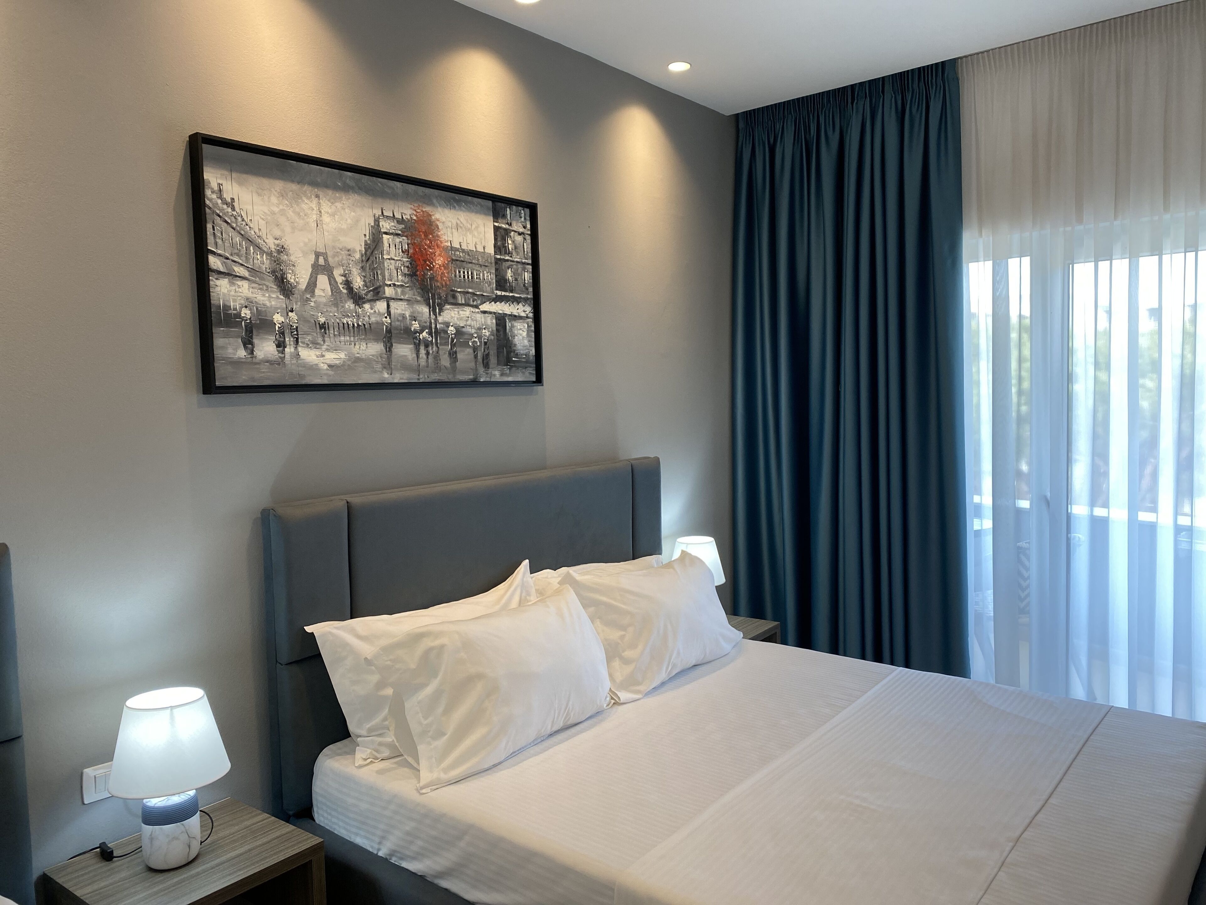 A hotel room with a bed, a nightstand with a lamp, a framed picture on the wall, and a window with curtains.