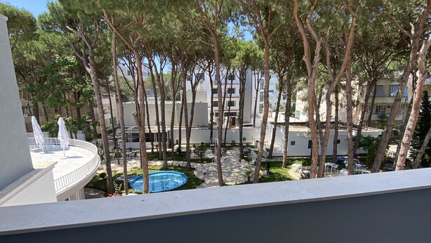 Un balcón con una mesa de mármol, una pequeña planta en maceta y vistas a una piscina rodeada de árboles y edificios.