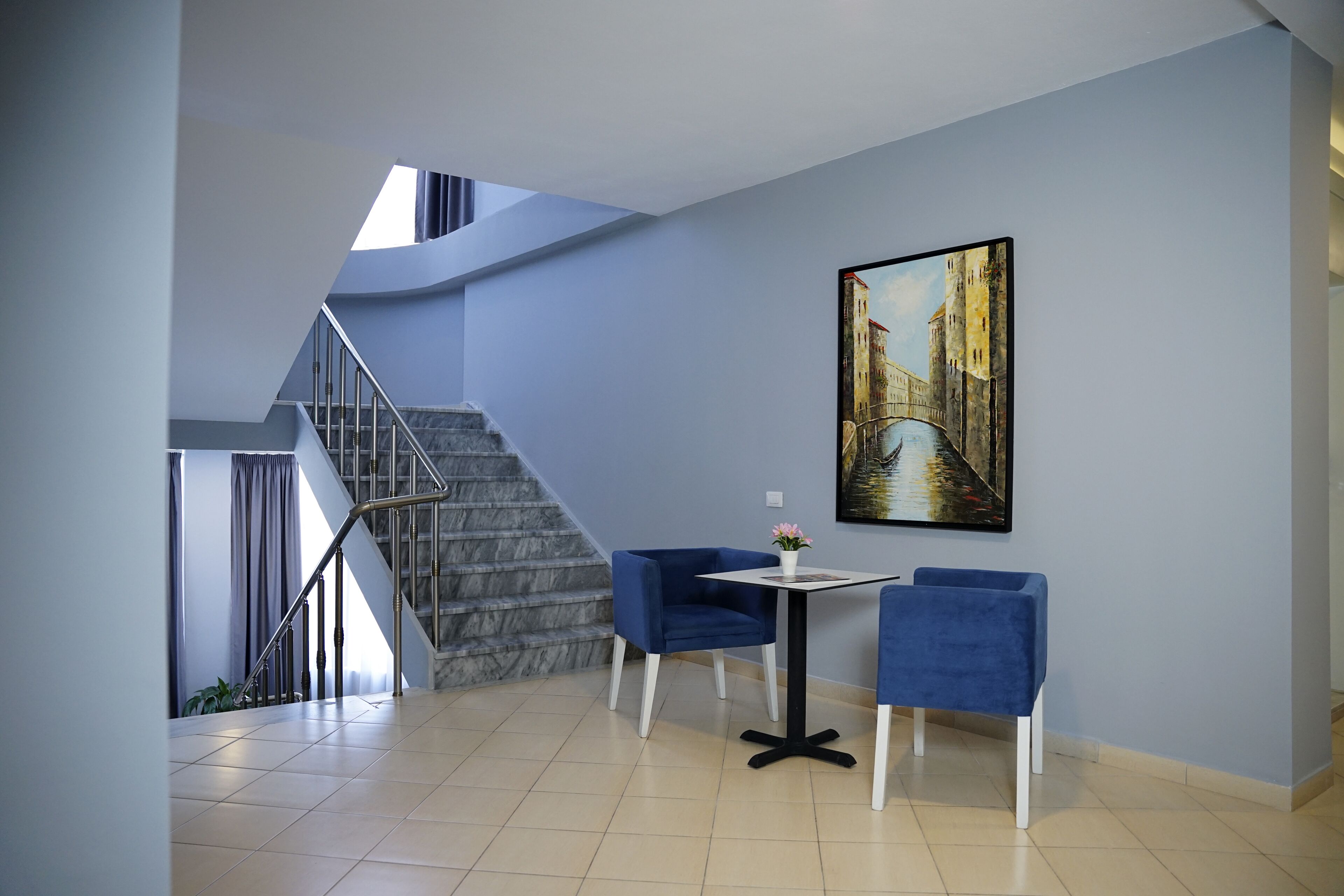 Um interior moderno com uma escada, duas cadeiras azuis, uma mesa pequena e um quadro grande na parede.