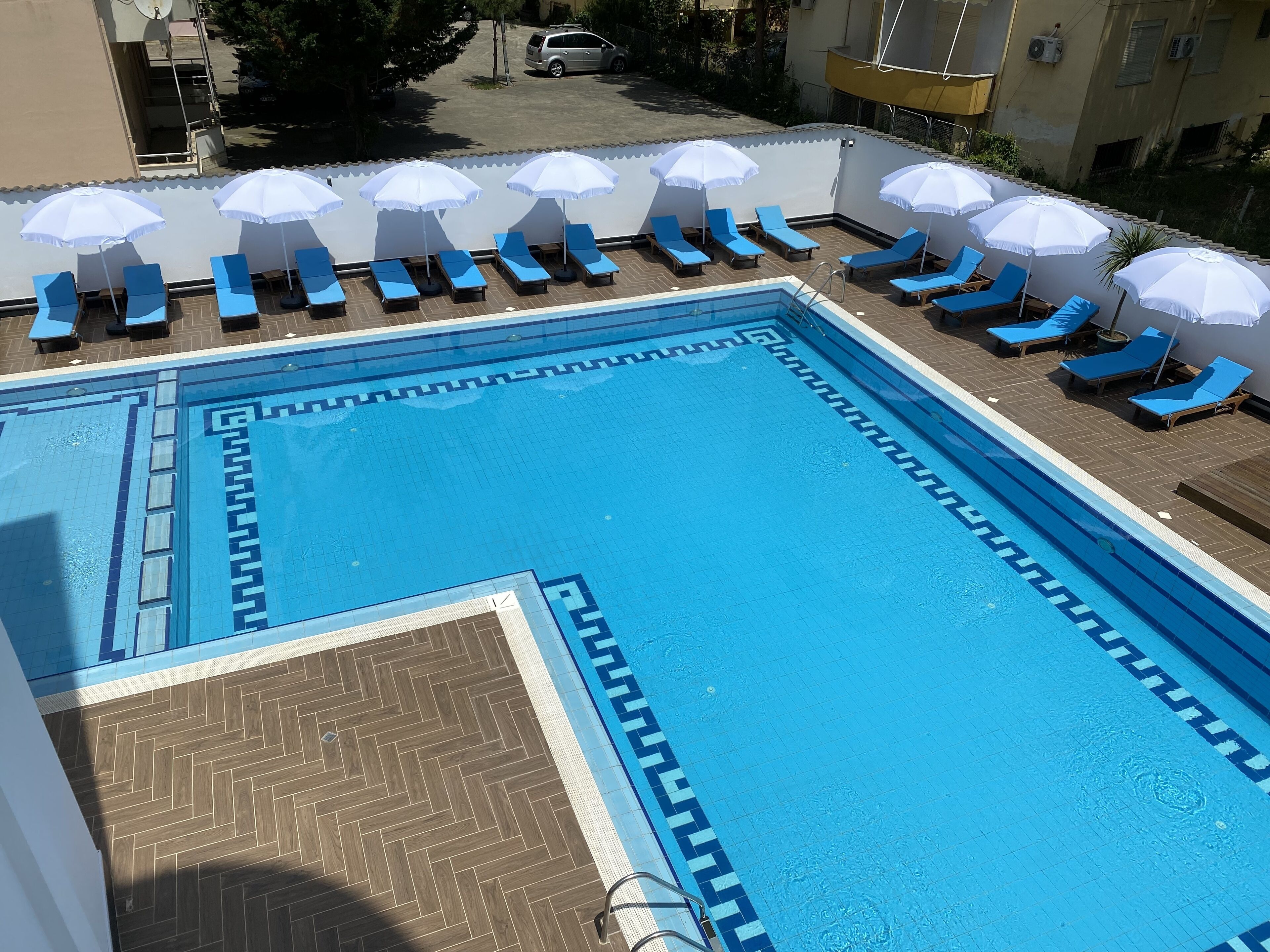 A swimming pool with blue tiles and a herringbone pattern around the edges. There are blue lounge chairs and white umbrellas arranged around the pool.