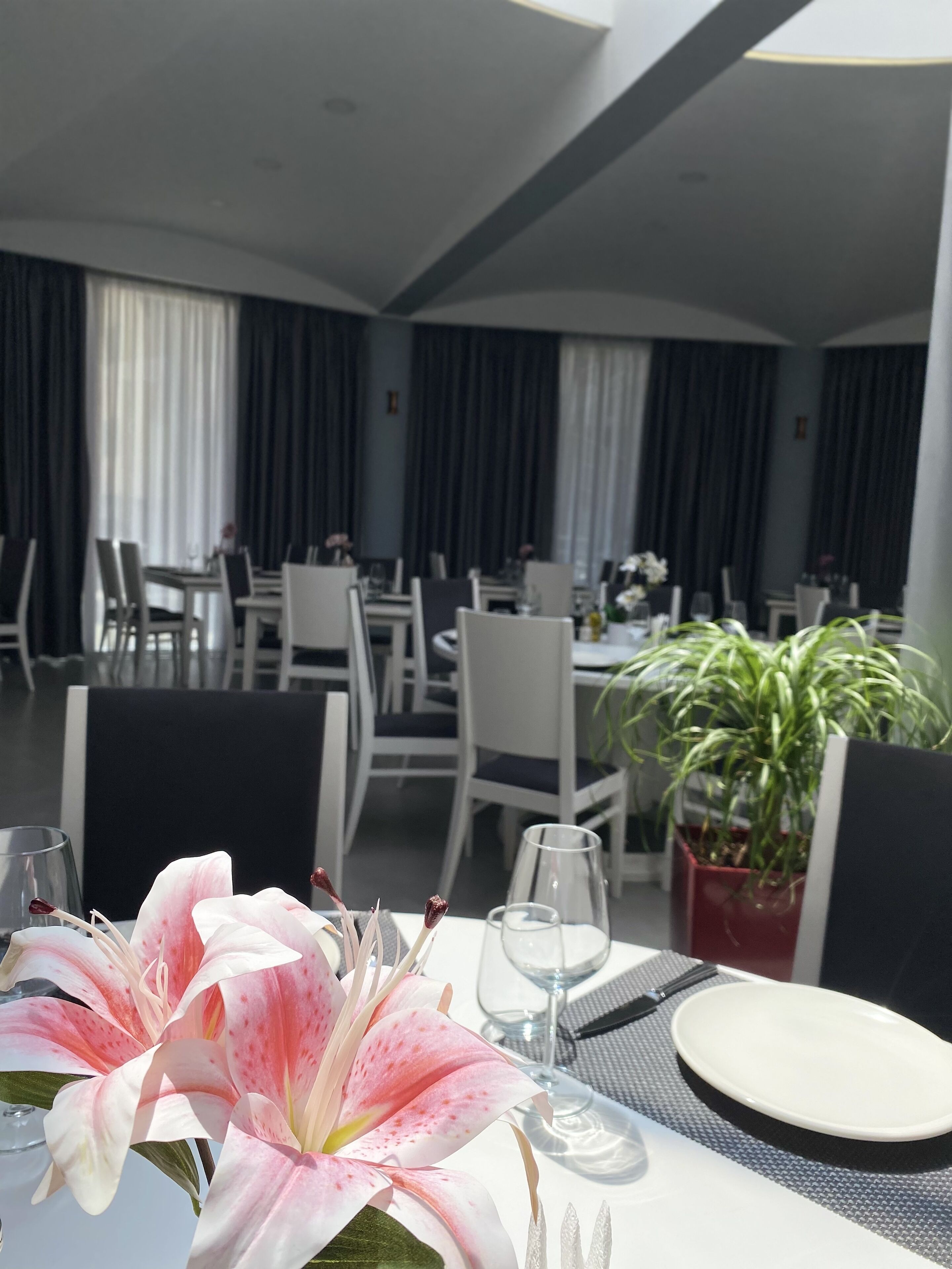 Un espace repas avec des tables préparées pour deux personnes, agrémenté de nappes blanches, de verres transparents et d’une plante en pot.