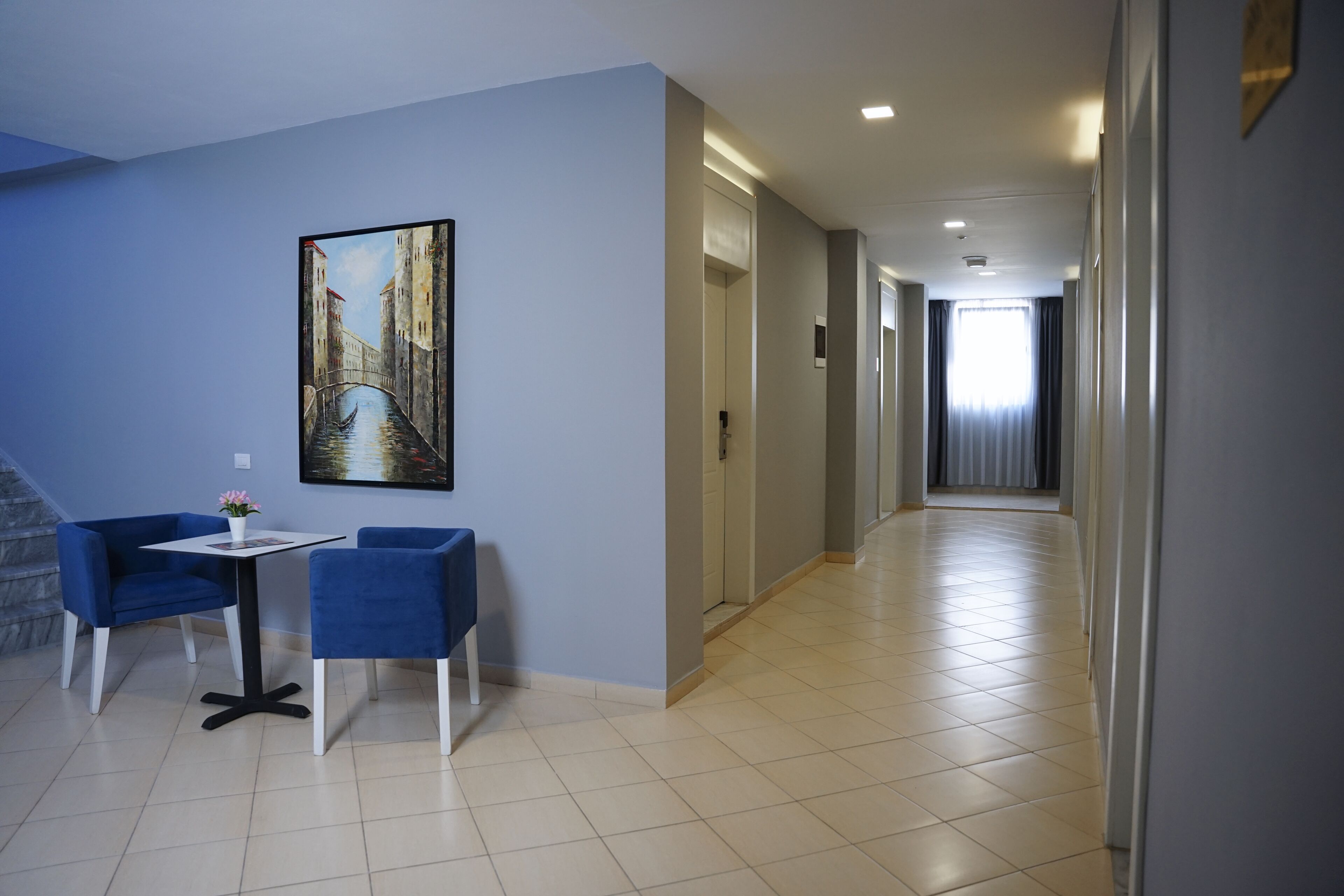 A hotel corridor with a painting of a canal, a small table with chairs, and a staircase to the left.