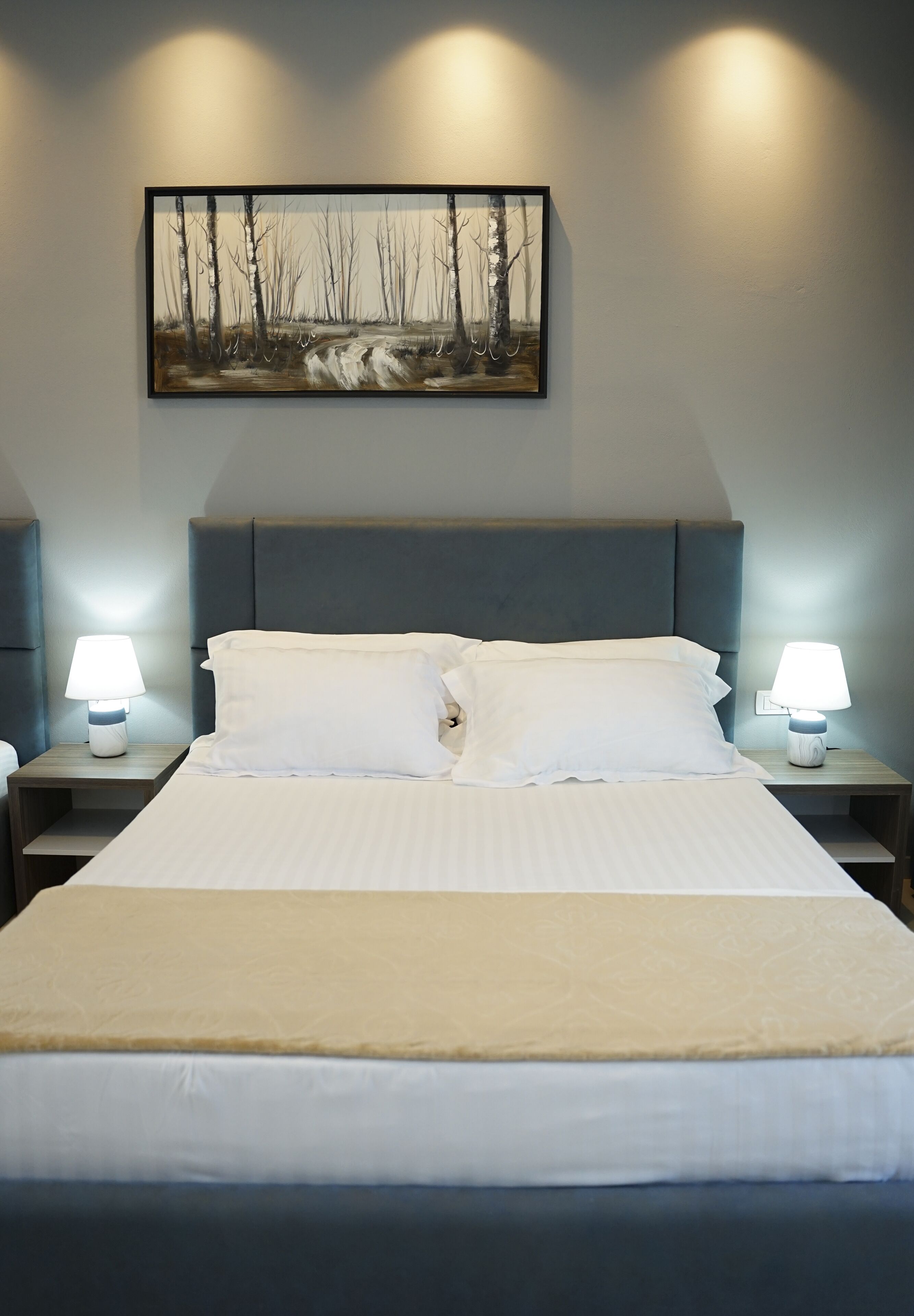 Een keurig opgemaakt bed met witte lakens, twee bedlampjes en een lijst met daarop een afbeelding van een bos.