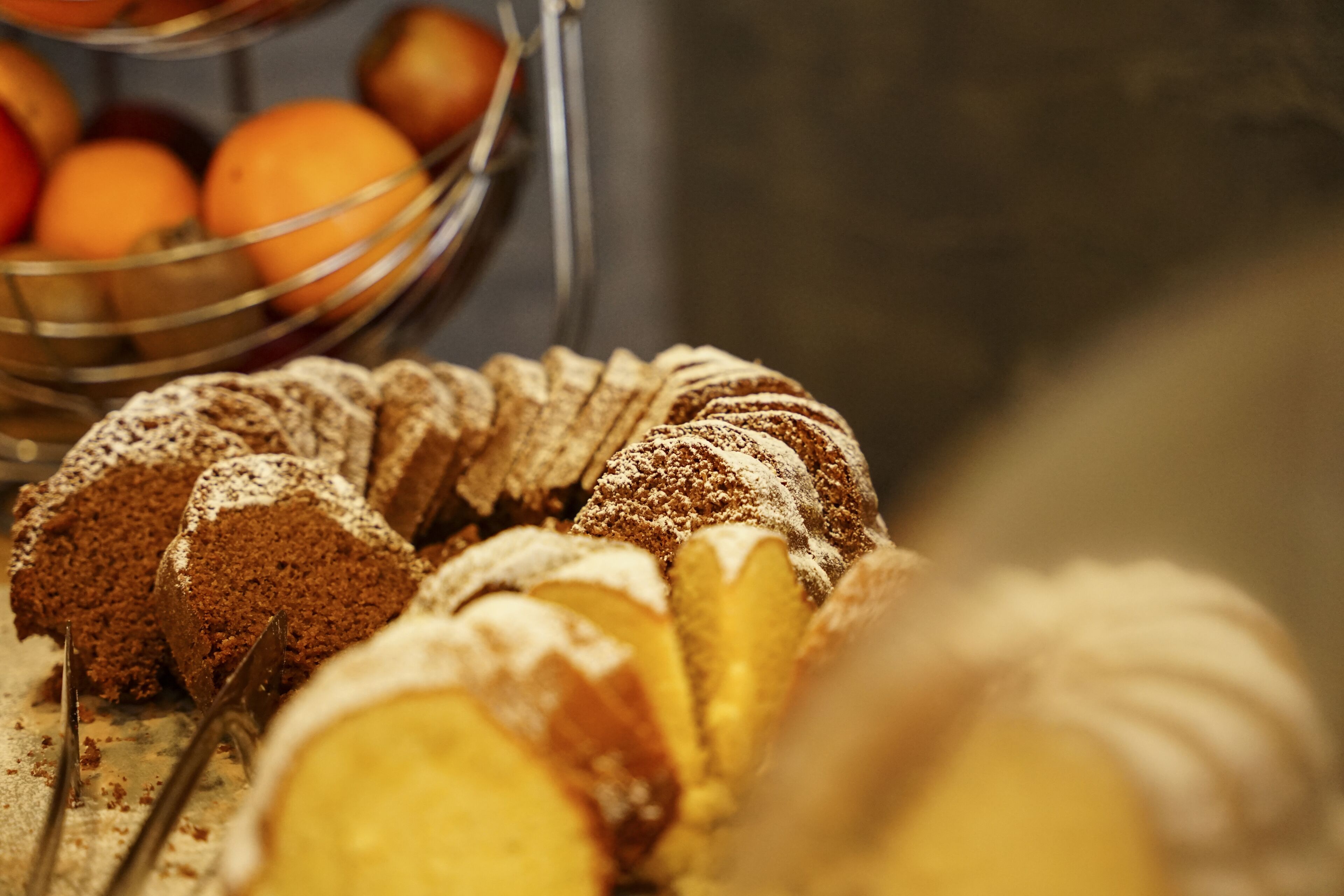 A close-up of a dessert with a dusting of powdered sugar, a metal serving tray, and a blurred background with oranges.