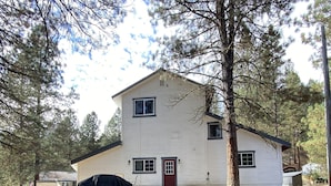 A two-story house with a red door and white siding, surrounded by pine trees and a covered grill area. - Cozy home with HOT TUB, River View & Fire Pit! (Garden Valley)