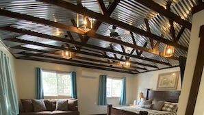 A spacious room with a large wooden floor, a cozy sofa, a checkered bed, and a rustic ceiling with exposed beams and hanging lights. - Cozy home with HOT TUB, River View & Fire Pit! (Garden Valley)