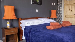 Une chambre à coucher avec un lit en bois, du linge de lit blanc, une couverture bleue, deux lampes orange et une table de chevet en bois sur laquelle se trouve une bouteille.