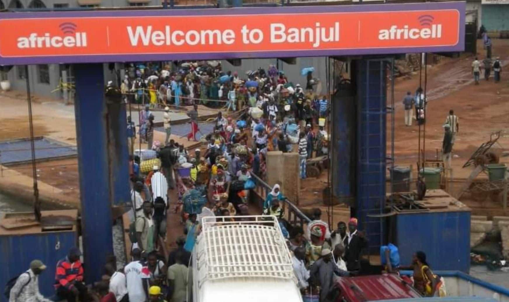 Escena concurrida en una estación de autobuses, con un cartel que dice 'Bienvenidos a Banjul' y logotipos de 'africell'.