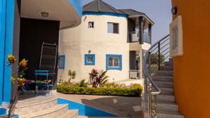 Un bâtiment à deux étages, à la façade bleue et beige, avec un escalier sur le côté droit et un petit espace extérieur aménagé avec des chaises bleues et des plantes.