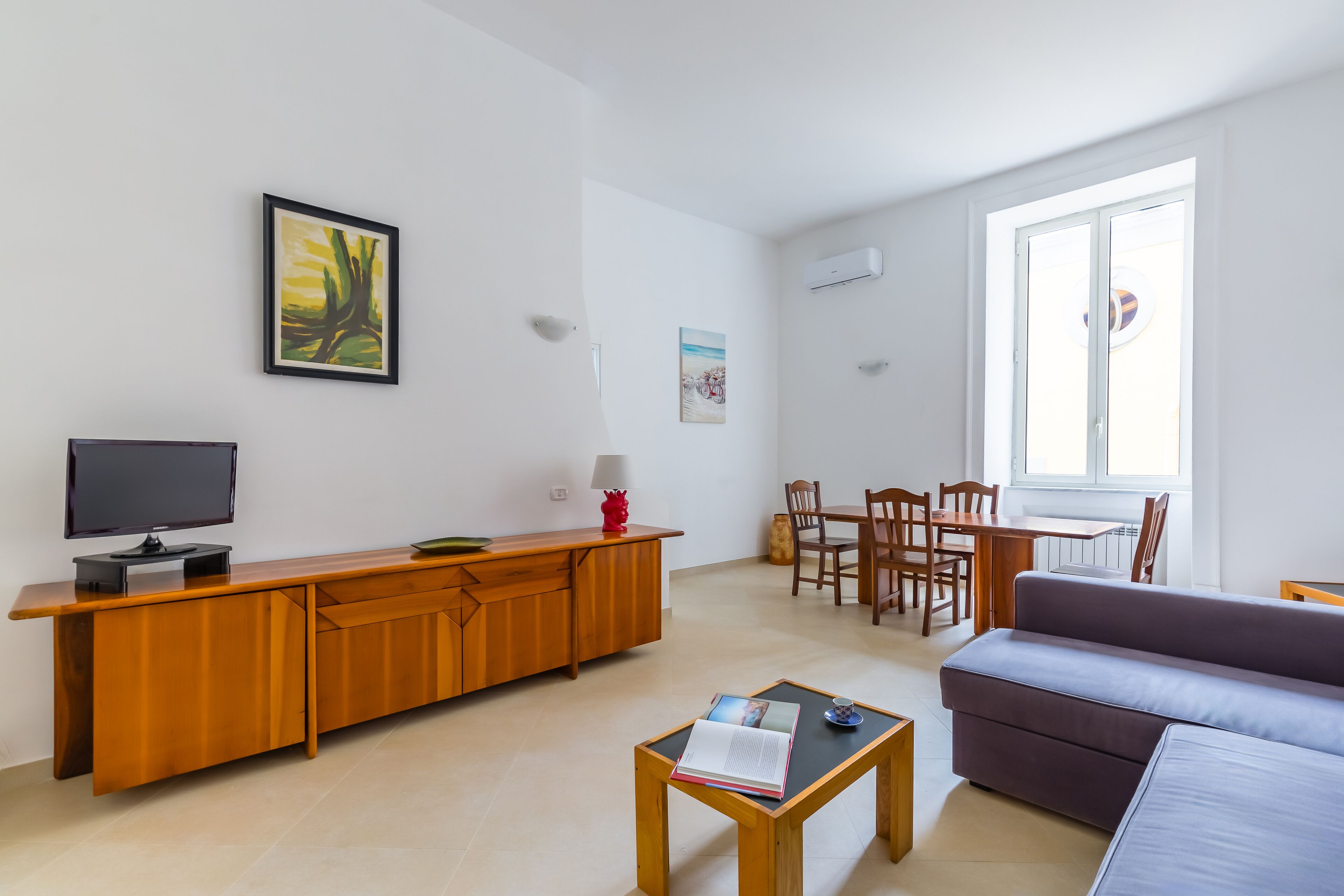 Un salon moderne comprenant un canapé, une table basse avec un livre, un téléviseur posé sur un meuble en bois, un coin repas avec des chaises et une table, et une grande fenêtre offrant une vue dégagée.