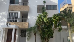 Un bâtiment moderne à deux étages, doté d’un toit plat, d’un balcon au deuxième étage et d’un petit jardin agrémenté de palmiers.