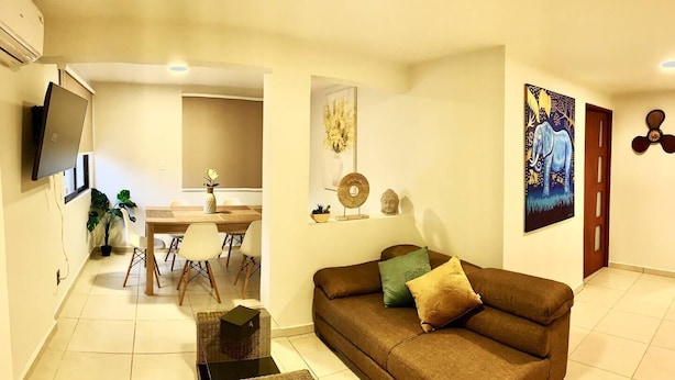 Un salon avec un canapé marron, une table à manger avec des chaises, un téléviseur fixé au mur et un tableau accroché au mur.