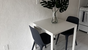 A dining area with a white table and two black chairs, a large green plant in a vase, and a painting of boats on the wall.