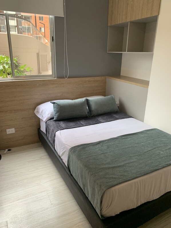 Quarto com cama, janela, cabeceira de madeira e prateleira.