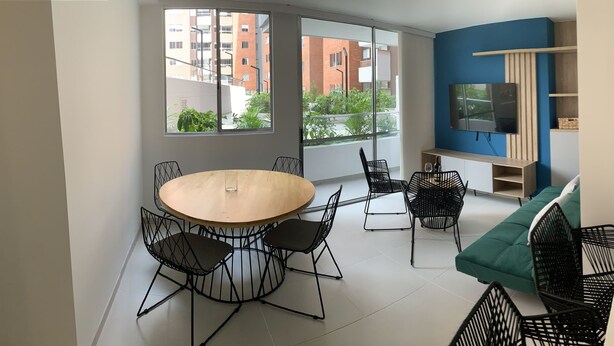 A modern apartment with a dining area featuring a round wooden table and black wireframe chairs, a living area with a TV and built-in shelves, and a kitchen with a sink and cabinets. - 3BR magical aparment in the heart of Medellin - Poblado  (Medellín)