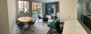Eine moderne Wohnung mit Essbereich, inklusive rundem Holztisch und schwarzen Stuhlgestellen, einem Wohnbereich mit Fernseher und Einbauschränken sowie einer Küche mit Spüle und Schränken.