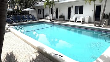 A rectangular pool with a 6 ft depth marker, surrounded by lounge chairs and palm trees, with a building in the background.