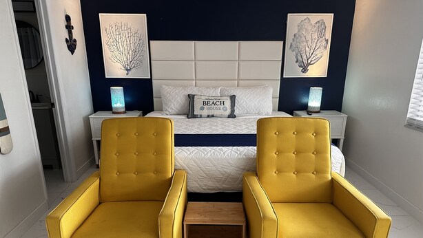 Een slaapkamer met een tweepersoonsbed, twee nachtkastjes, twee gele fauteuils en kunstwerken aan de muur.