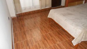 Una habitación con piso de madera, una cama con sábana blanca, un armario de madera y una ventana con cortinas traslúcidas.