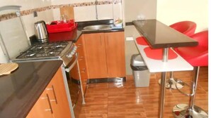 Cocina compacta con estufa a gas, armarios de madera, pileta de acero inoxidable y un pequeño bote de basura.